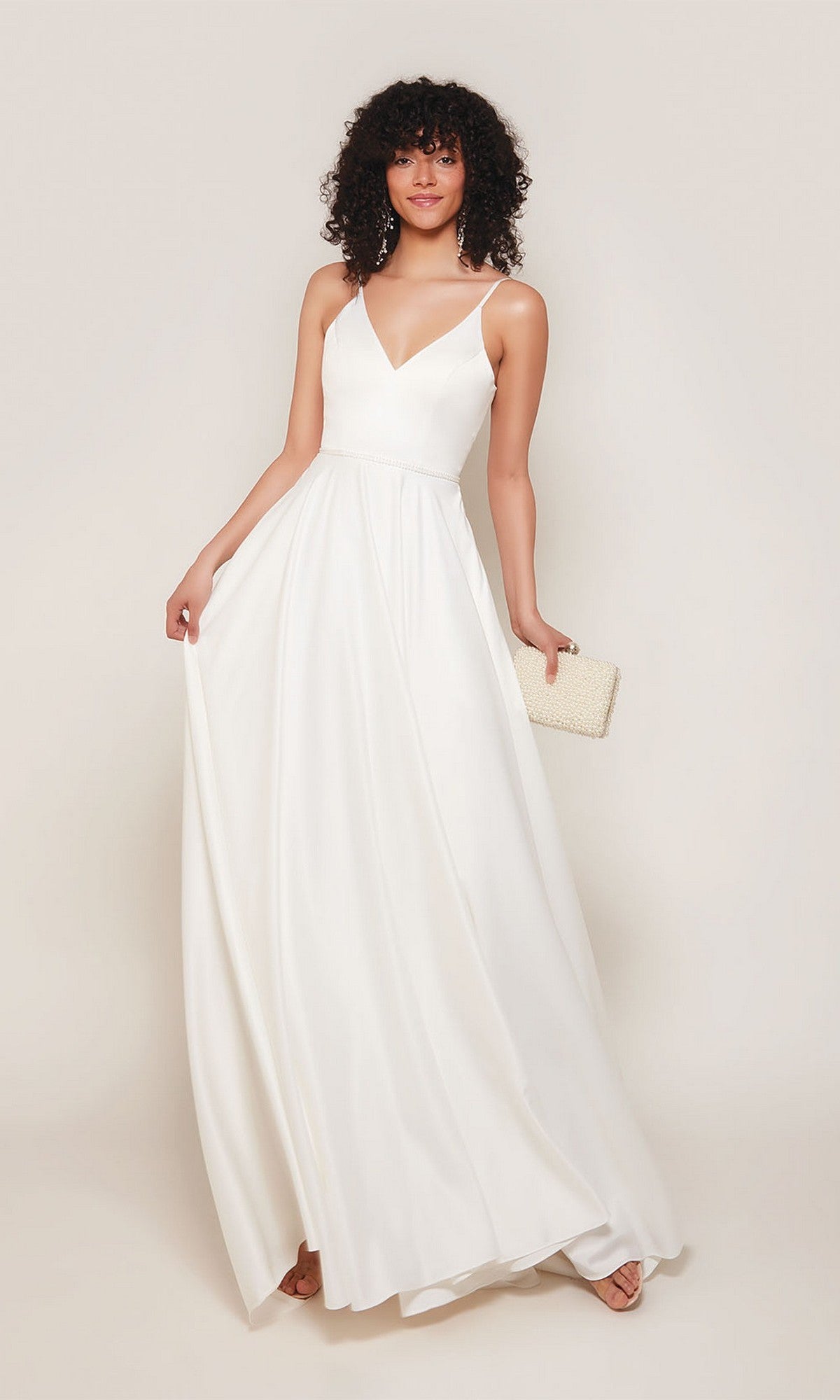 A woman wearing the Alyce 7089 long formal dress in diamond white with thin straps stands against a plain background, holding a textured beige clutch and smiling.